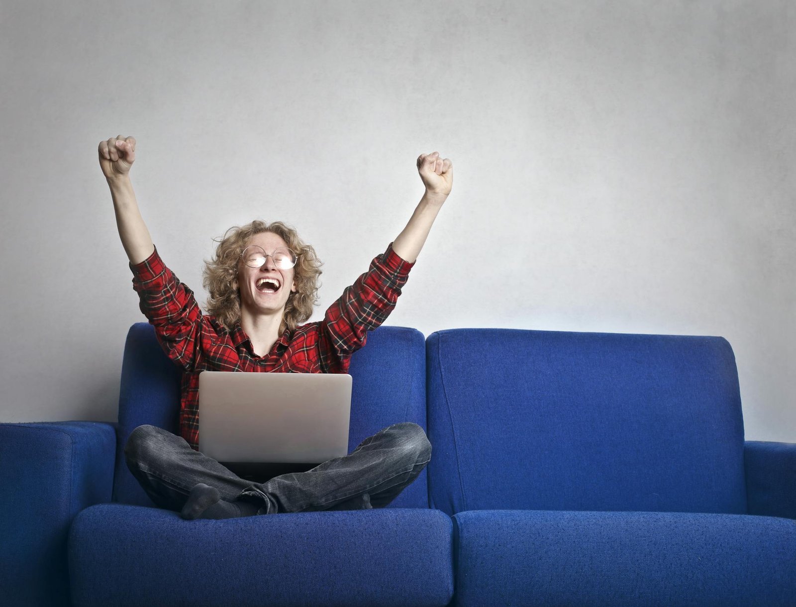 Thrilled individual celebrating success while using a laptop on a blue sofa indoors.